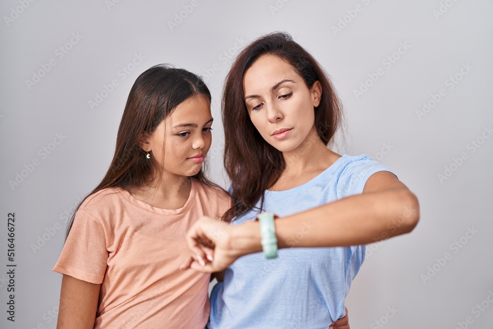 Young mother and daughter standing over white background checking the time on wrist watch, relaxed and confident Young mother and daughter standing over white background checking the time on wrist watch, relaxed and confident