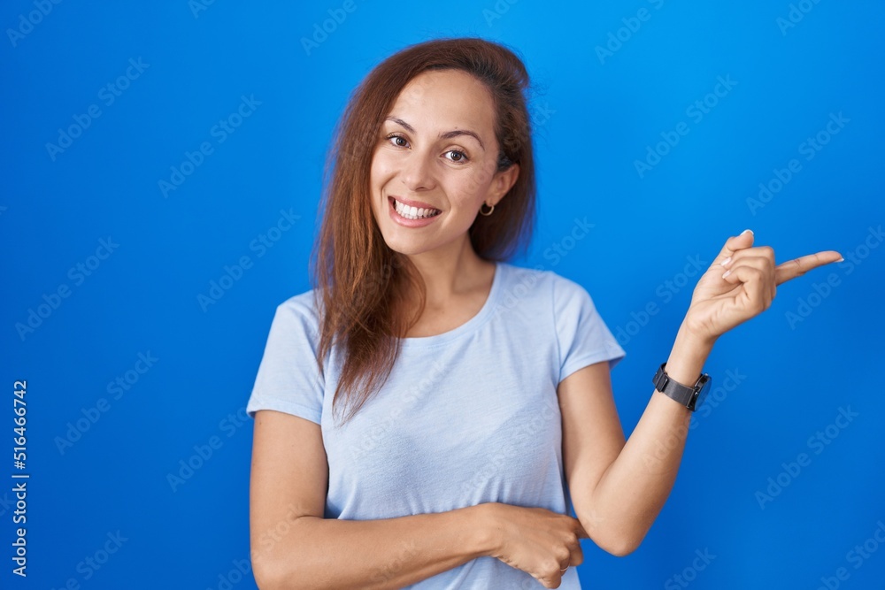 Brunette woman standing over blue background with a big smile on face, pointing with hand finger to the side looking at the camera. Brunette woman standing over blue background with a big smile on face, pointing with hand finger to the side looking at the camera.