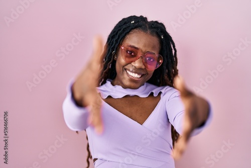 African woman with braided hair standing over pink background looking at the camera smiling with open arms for hug. cheerful expression embracing happiness.
