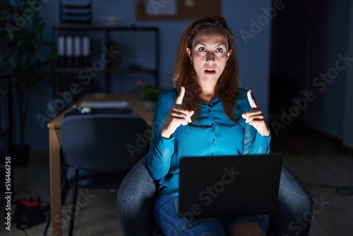 Brunette woman working at the office at night amazed and surprised looking up and pointing with fingers and raised arms.