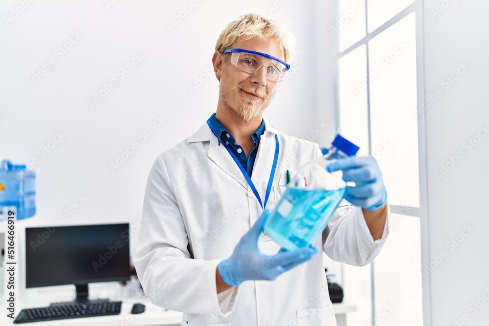 Young caucasian man wearing scientist uniform holding bottle with liquid at laboratory