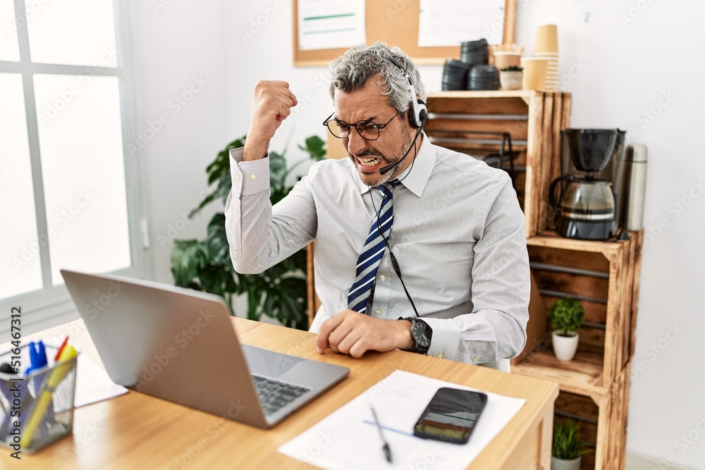 Middle age hispanic business man working at the office wearing operator headset angry and mad raising fist frustrated and furious while shouting with anger. rage and aggressive concept. Middle age hispanic business man working at the office wearing operator headset angry and mad raising fist frustrated and furious while shouting with anger. rage and aggressive concept.
