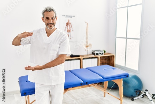 Middle age hispanic therapist man working at pain recovery clinic gesturing with hands showing big and large size sign  measure symbol. smiling looking at the camera. measuring concept.