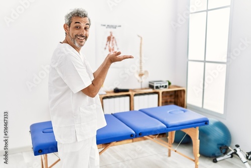 Middle age hispanic therapist man working at pain recovery clinic pointing aside with hands open palms showing copy space, presenting advertisement smiling excited happy