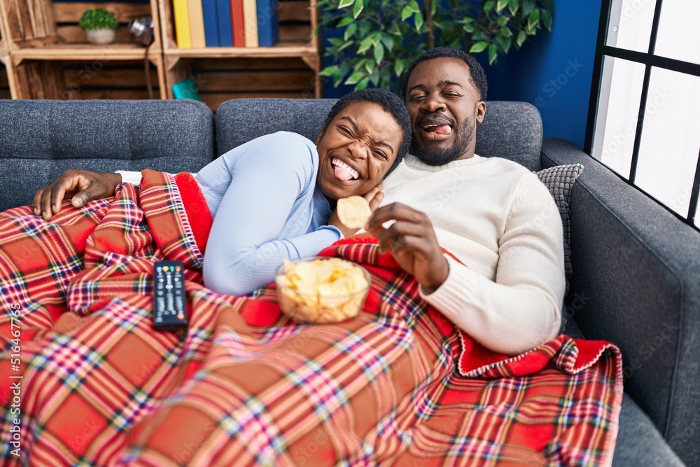 Young african american couple sitting on the sofa watching tv sticking tongue out happy with funny expression. - obrazy, fototapety, plakaty 