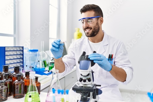 Young hispanic man wearing scientist uniform using microscope at laboratory