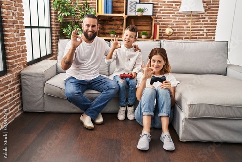 Family of three playing video game sitting on the sofa doing ok sign with fingers, smiling friendly gesturing excellent symbol