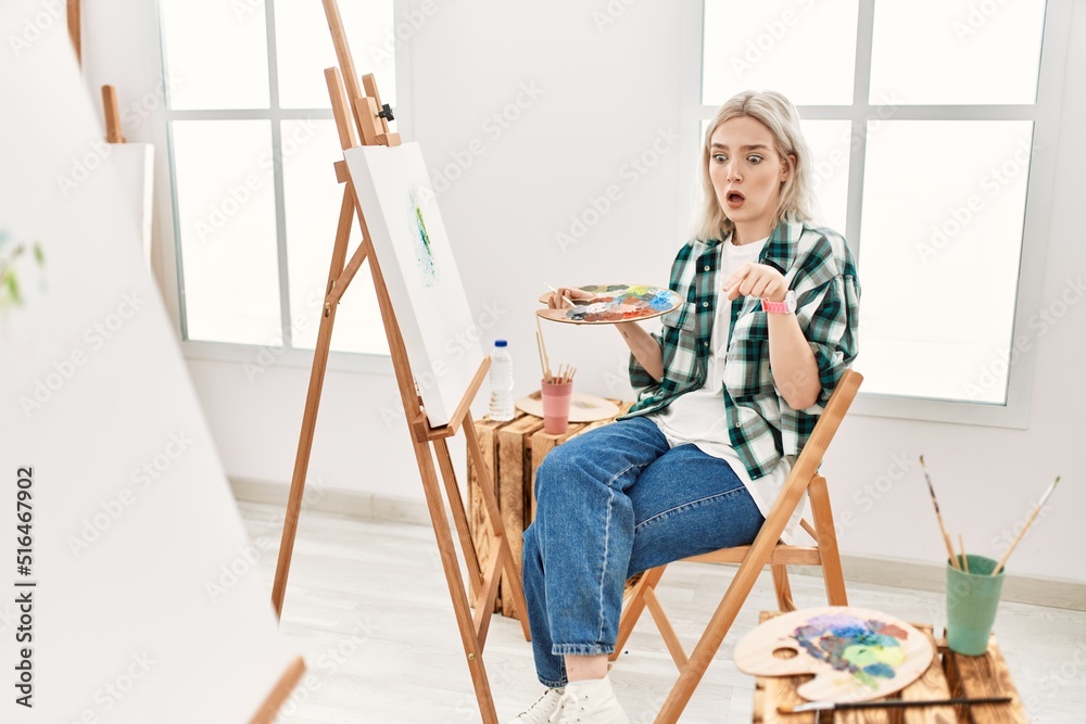 Young artist woman painting on canvas at art studio pointing down with fingers showing advertisement, surprised face and open mouth Young artist woman painting on canvas at art studio pointing down with fingers showing advertisement, surprised face and open mouth