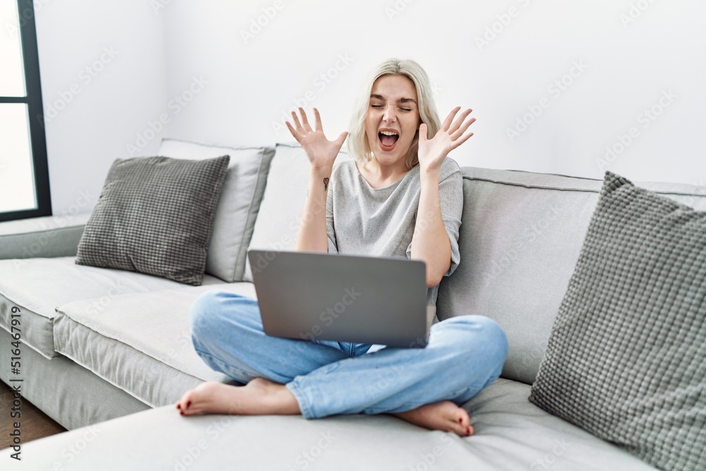 Young caucasian woman using laptop at home sitting on the sofa celebrating mad and crazy for success with arms raised and closed eyes screaming excited. winner concept Young caucasian woman using laptop at home sitting on the sofa celebrating mad and crazy for success with arms raised and closed eyes screaming excited. winner concept