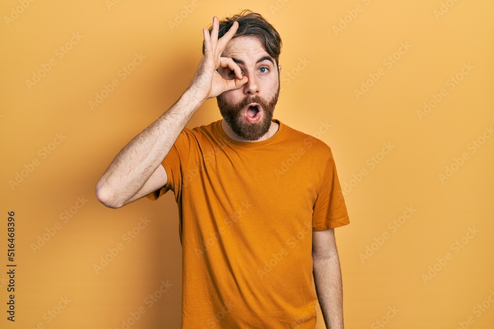 Caucasian man with beard wearing casual yellow t shirt doing ok gesture shocked with surprised face, eye looking through fingers. unbelieving expression. Caucasian man with beard wearing casual yellow t shirt doing ok gesture shocked with surprised face, eye looking through fingers. unbelieving expression.