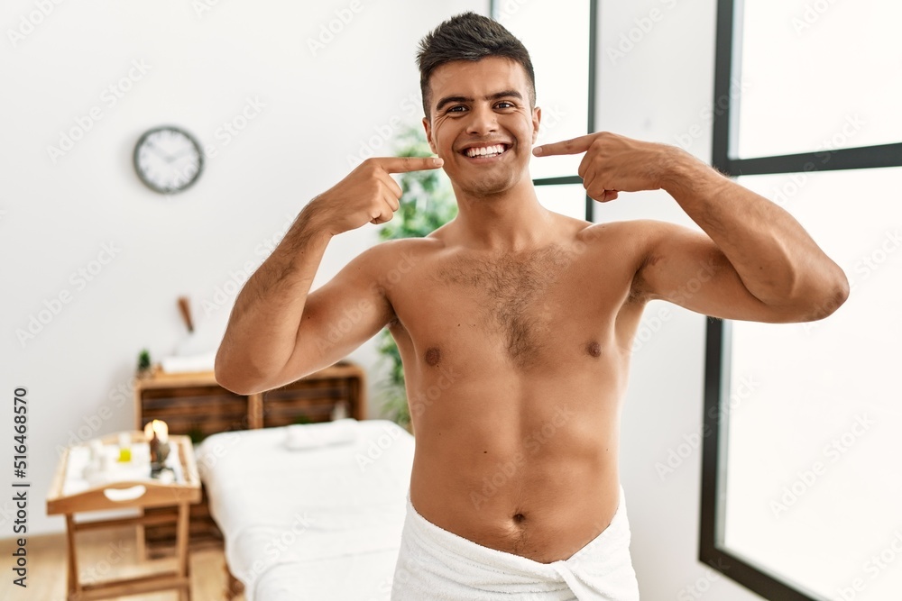 Young hispanic man standing shirtless at spa center smiling cheerful showing and pointing with fingers teeth and mouth. dental health concept.