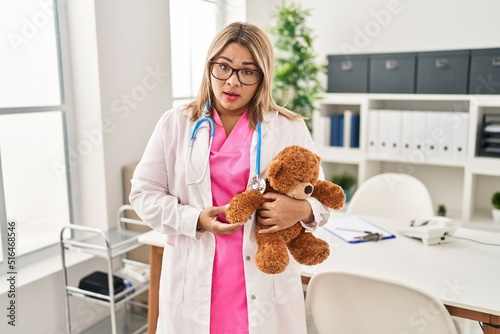 Young hispanic doctor woman holding teddy bear scared and amazed with open mouth for surprise, disbelief face