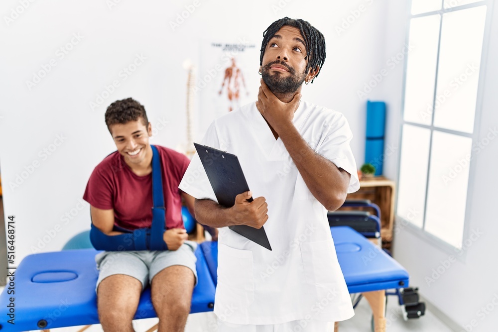 Fototapeta premium Young hispanic man working at pain recovery clinic with a man with broken arm touching painful neck, sore throat for flu, clod and infection