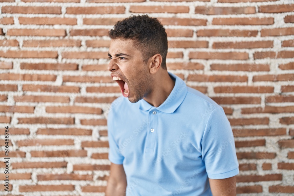Brazilian young man standing over brick wall angry and mad screaming frustrated and furious, shouting with anger. rage and aggressive concept. Brazilian young man standing over brick wall angry and mad screaming frustrated and furious, shouting with anger. rage and aggressive concept.