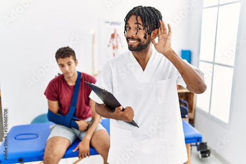 Young hispanic man working at pain recovery clinic with a man with broken arm smiling with hand over ear listening an hearing to rumor or gossip. deafness concept.