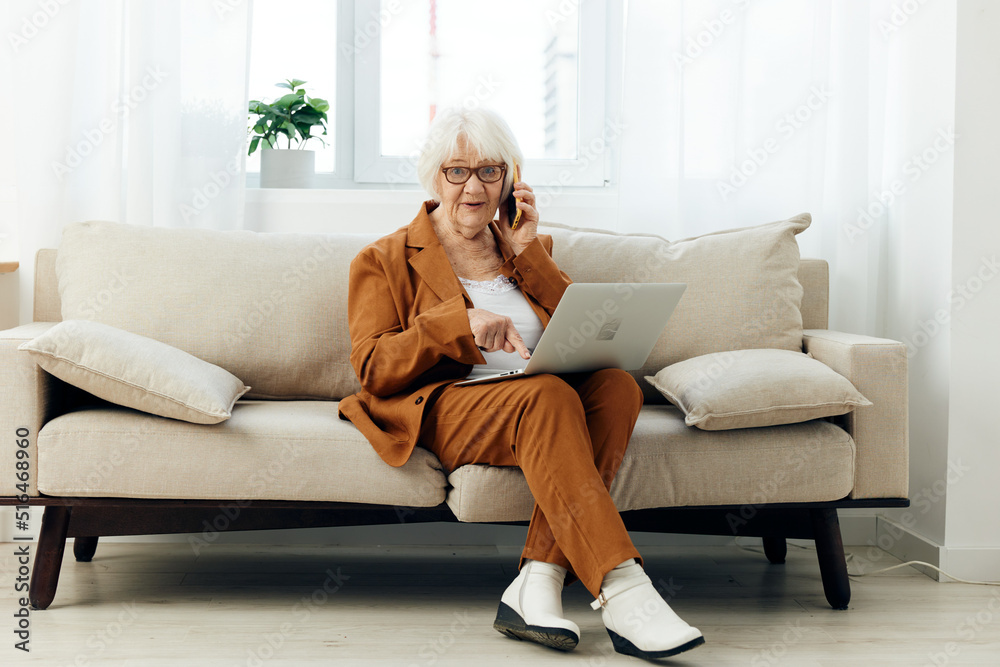 a joyful elderly businesswoman is sitting on a cozy sofa in a bright apartment holding a laptop on her lap and gesturing with her hand while solving work issues on the phone