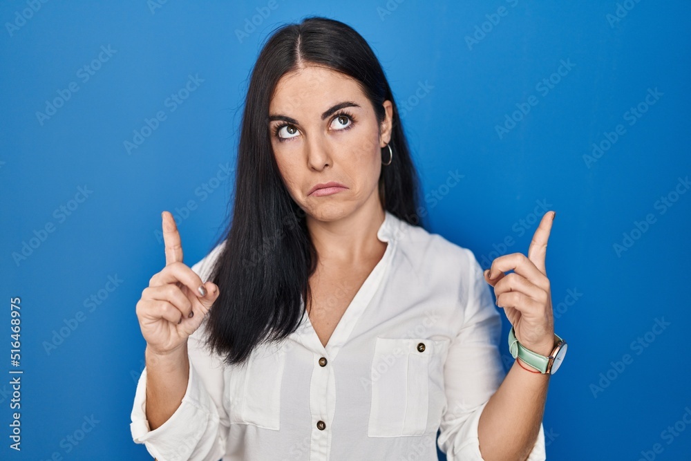 Young hispanic woman standing over blue background pointing up looking sad and upset, indicating direction with fingers, unhappy and depressed. Young hispanic woman standing over blue background pointing up looking sad and upset, indicating direction with fingers, unhappy and depressed.
