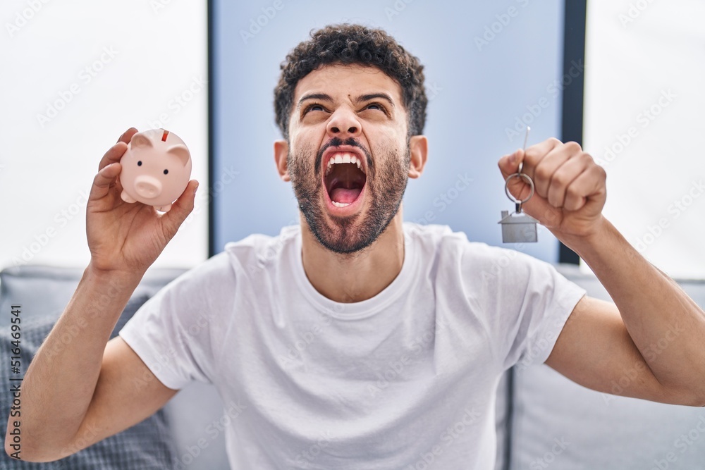 Arab man holding piggy bank and house keys angry and mad screaming frustrated and furious, shouting with anger looking up.
