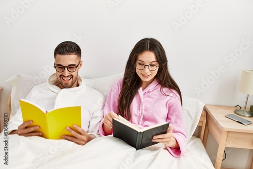 Young hispanic couple reading book lying in bed at home.