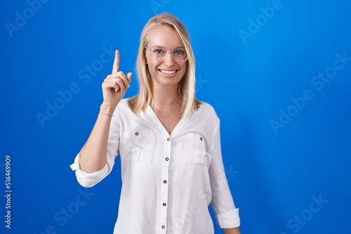 Young caucasian woman standing over blue background pointing finger up with successful idea. exited and happy. number one.