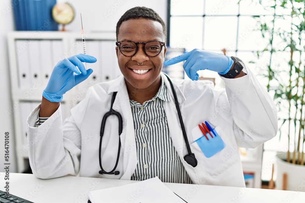 Young african doctor man holding syringe at the hospital smiling cheerful showing and pointing with fingers teeth and mouth. dental health concept.