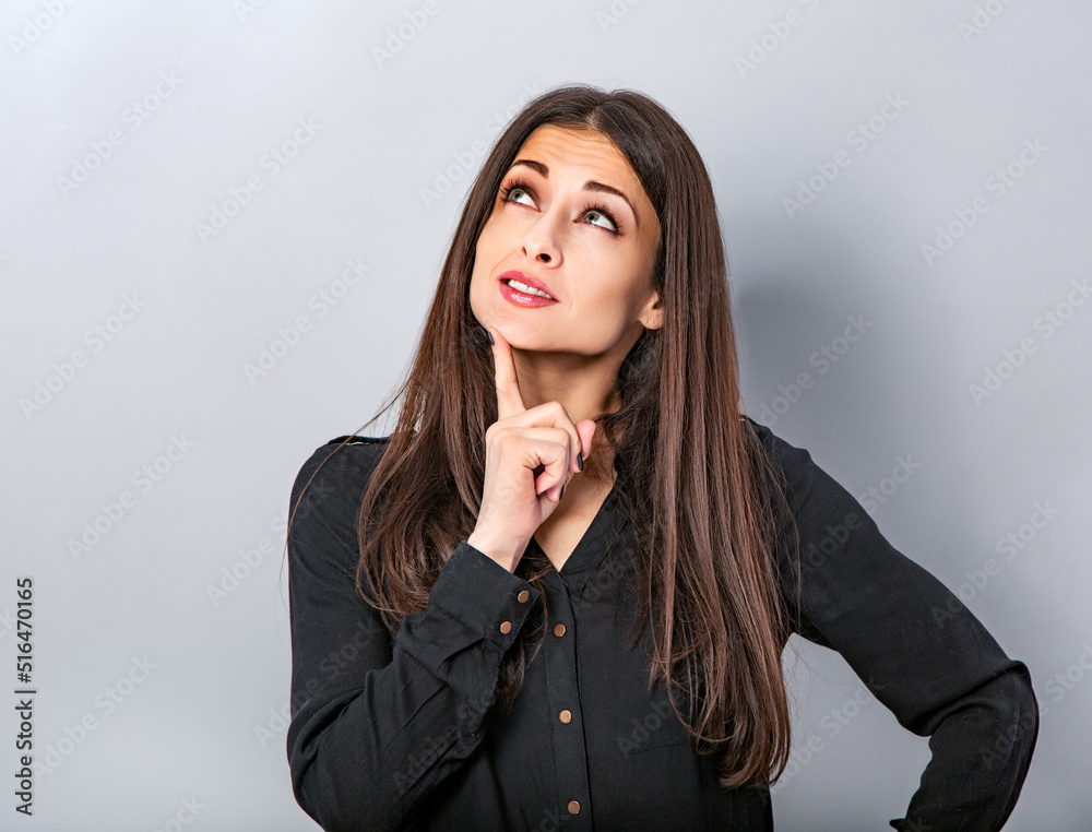 Beautiful young business serious woman thinking and looking up with finger under the face on blue grey background with empty copy space. Portrait