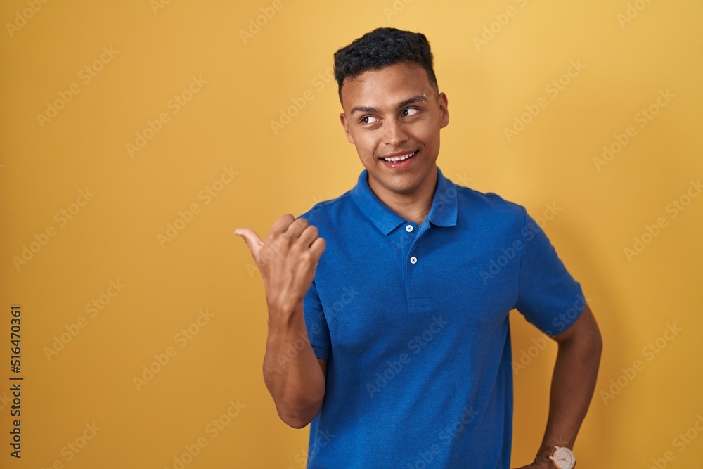 Young hispanic man standing over yellow background smiling with happy face looking and pointing to the side with thumb up. Young hispanic man standing over yellow background smiling with happy face looking and pointing to the side with thumb up.