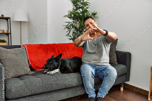 Young latin man and dog sitting on the sofa at home smiling in love doing heart symbol shape with hands. romantic concept. © Krakenimages.com