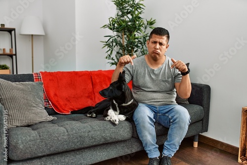 Young latin man and dog sitting on the sofa at home pointing down looking sad and upset, indicating direction with fingers, unhappy and depressed. © Krakenimages.com