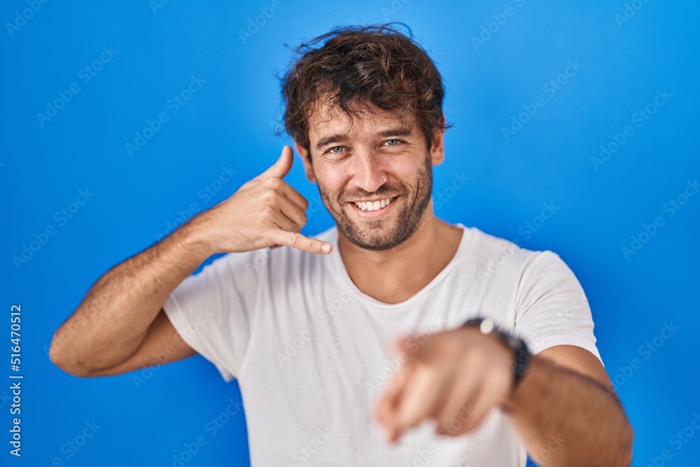 Hispanic young man standing over blue background smiling doing talking on the telephone gesture and pointing to you. call me. Hispanic young man standing over blue background smiling doing talking on the telephone gesture and pointing to you. call me.
