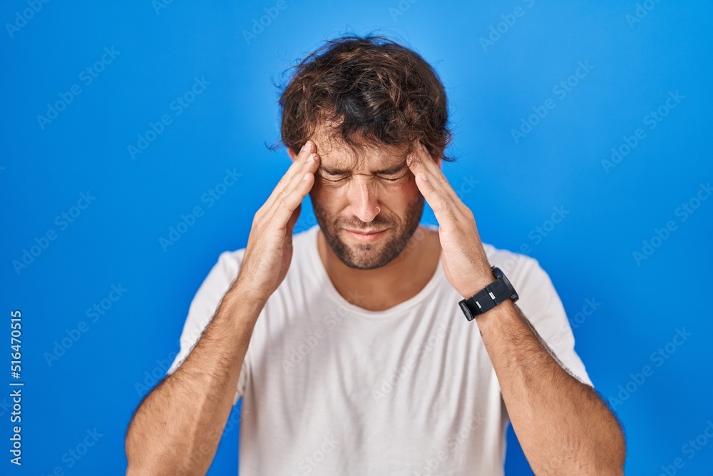 Hispanic young man standing over blue background with hand on head, headache because stress. suffering migraine. Hispanic young man standing over blue background with hand on head, headache because stress. suffering migraine.