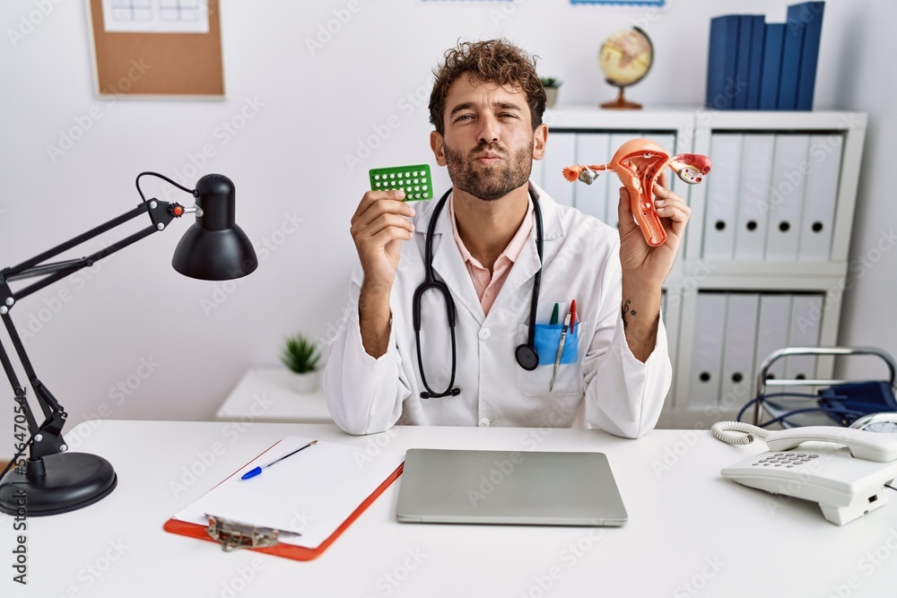 Obraz premium Young hispanic doctor man holding female genital organ and birth control pills looking at the camera blowing a kiss being lovely and sexy. love expression.