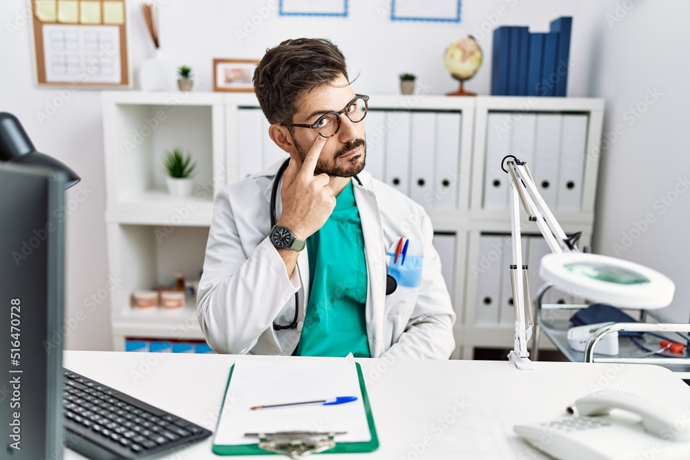 Young man with beard wearing doctor uniform and stethoscope at the clinic pointing to the eye watching you gesture, suspicious expression Young man with beard wearing doctor uniform and stethoscope at the clinic pointing to the eye watching you gesture, suspicious expression