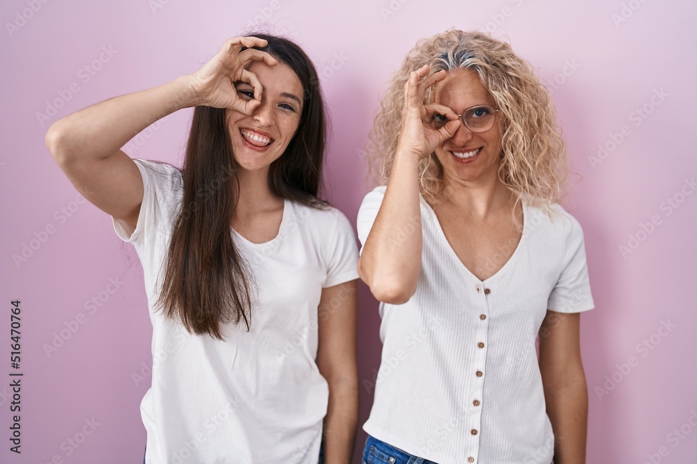 Mother and daughter standing together over pink background doing ok gesture with hand smiling, eye looking through fingers with happy face. Mother and daughter standing together over pink background doing ok gesture with hand smiling, eye looking through fingers with happy face.