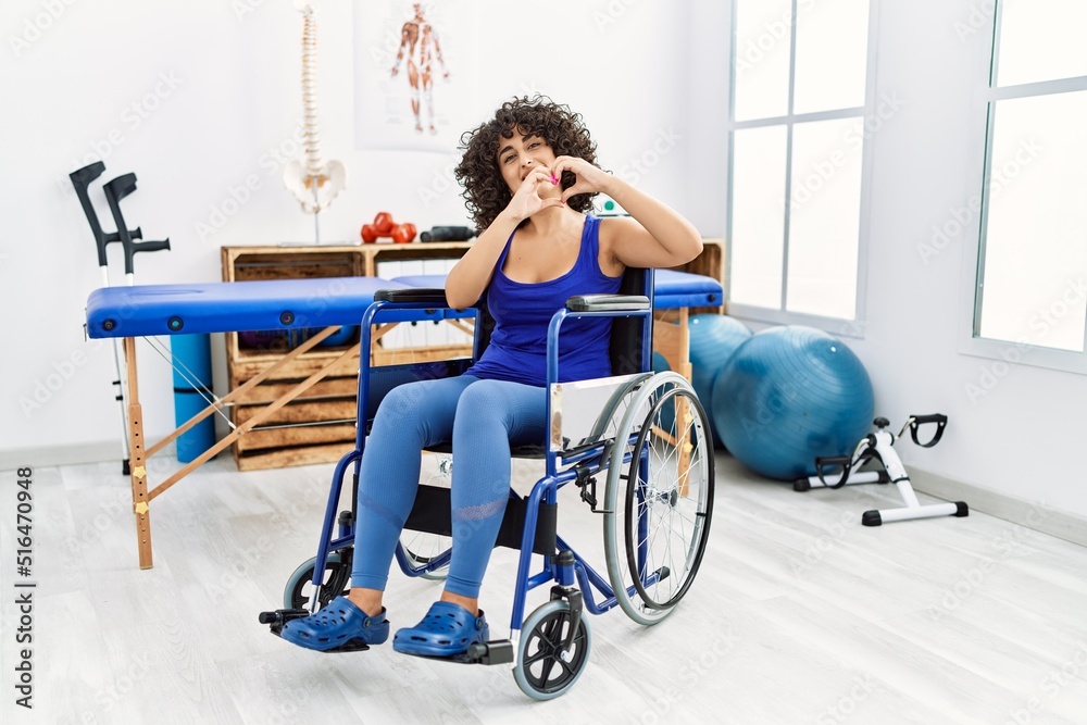 Obraz premium Young middle eastern woman sitting on wheelchair at physiotherapy clinic smiling in love doing heart symbol shape with hands. romantic concept.