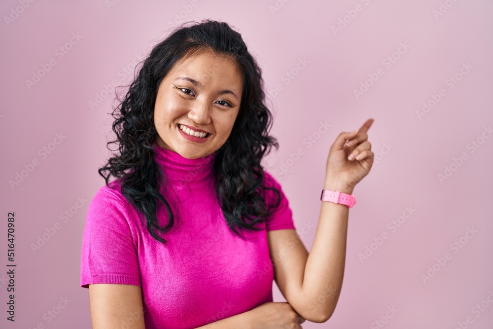 Young asian woman standing over pink background with a big smile on face, pointing with hand finger to the side looking at the camera. Young asian woman standing over pink background with a big smile on face, pointing with hand finger to the side looking at the camera.