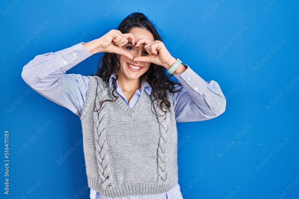 Young brunette woman standing over blue background doing heart shape with hand and fingers smiling looking through sign Young brunette woman standing over blue background doing heart shape with hand and fingers smiling looking through sign