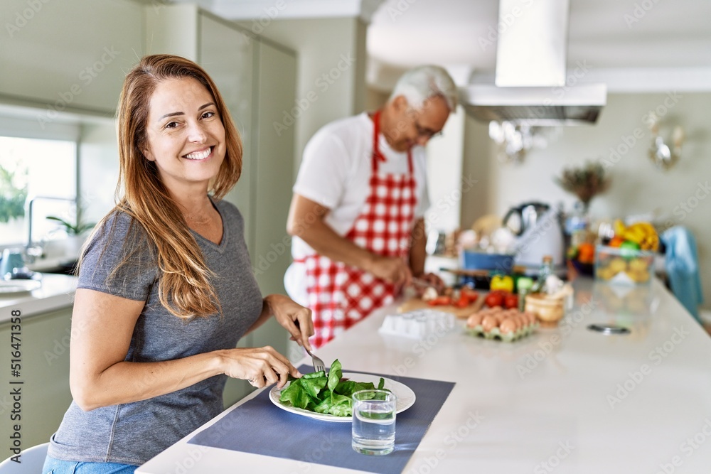 Middle age hispanic woman smiling happy eating salad while man cook at the kitchen. Middle age hispanic woman smiling happy eating salad while man cook at the kitchen.