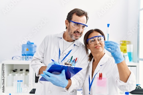 Middle age man and woman partners wearing scientist uniform writing on clipboard looking test tube at laboratory