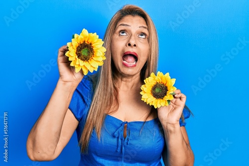 Middle age hispanic woman holding yellow sunflowers angry and mad screaming frustrated and furious  shouting with anger looking up.