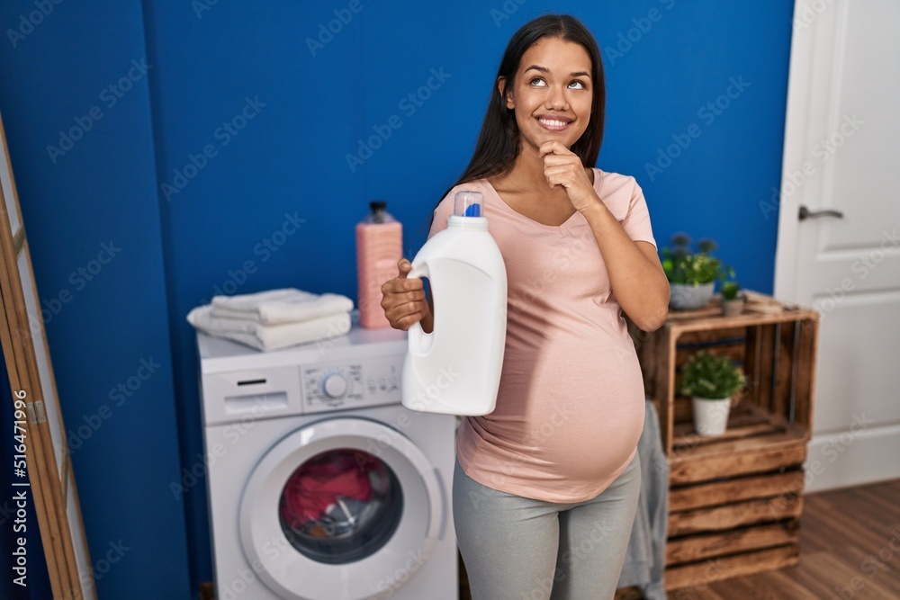 Young pregnant woman doing laundry holding detergent bottle serious face thinking about question with hand on chin, thoughtful about confusing idea Young pregnant woman doing laundry holding detergent bottle serious face thinking about question with hand on chin, thoughtful about confusing idea