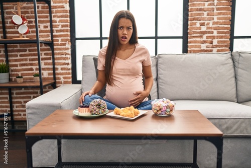 Young pregnant woman eating sweets at home scared and amazed with open mouth for surprise, disbelief face