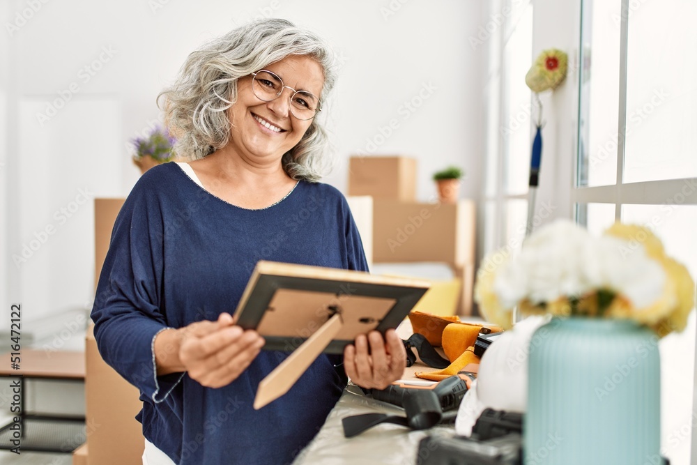 Middle age grey-haired woman smiling happy holding picture at new home. Middle age grey-haired woman smiling happy holding picture at new home.