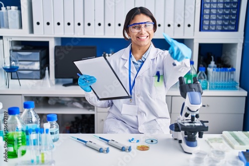 Hispanic young woman working at scientist laboratory approving doing positive gesture with hand  thumbs up smiling and happy for success. winner gesture.