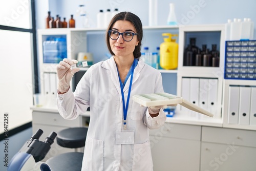 Young hispanic woman working at scientist laboratory with blood samples smiling looking to the side and staring away thinking.