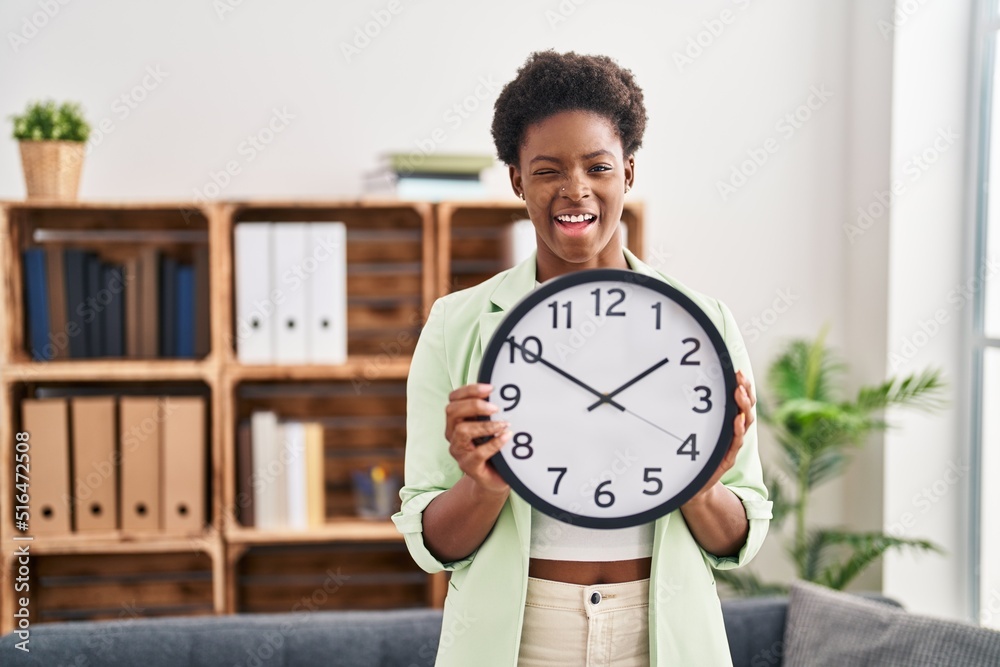 African american woman holding big clock winking looking at the camera with sexy expression, cheerful and happy face. African american woman holding big clock winking looking at the camera with sexy expression, cheerful and happy face.