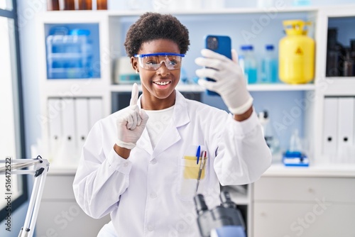 African american woman working at scientist laboratory doing video call with smartphone smiling with an idea or question pointing finger with happy face  number one