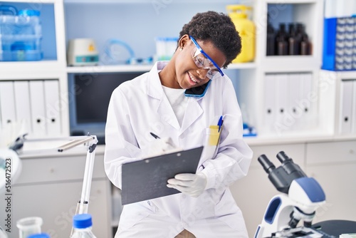 African american woman wearing scientist uniform talking on the smartphone write on clipboard at laboratory