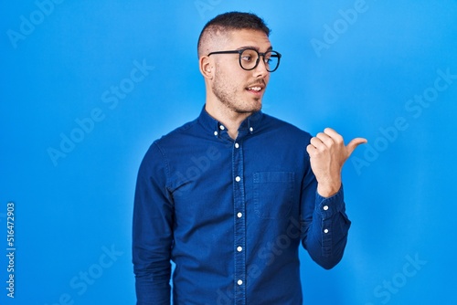 Young hispanic man wearing glasses over blue background smiling with happy face looking and pointing to the side with thumb up.