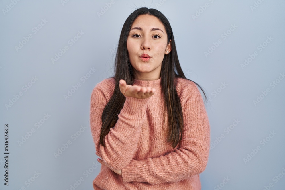 Fototapeta premium Young latin woman standing over blue background looking at the camera blowing a kiss with hand on air being lovely and sexy. love expression.